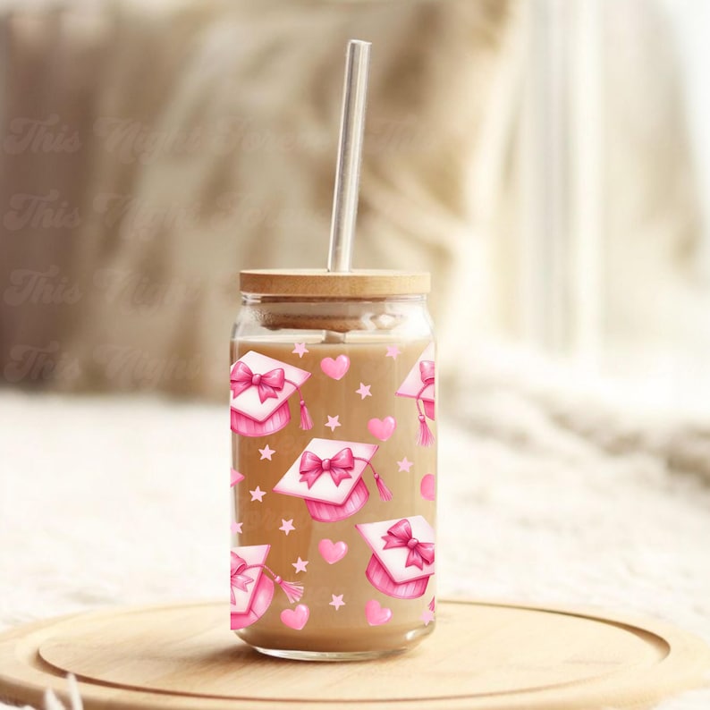 May include: A clear glass tumbler with a wooden lid and a clear straw. The tumbler is decorated with a pattern of pink graduation caps, bows, hearts, and stars. The tumbler is filled with a light brown liquid and sits on a wooden tray.