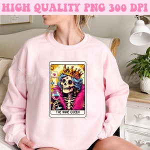 K&ouml;nnte beinhalten: Ein rosa Sweatshirt mit einem Grafikdruck einer Skelettk&ouml;nigin, die eine Krone tr&auml;gt und ein Glas Wein h&auml;lt. Die Grafik ist im Tarotkarten-Stil gehalten und tr&auml;gt den Schriftzug "The Wine Queen".