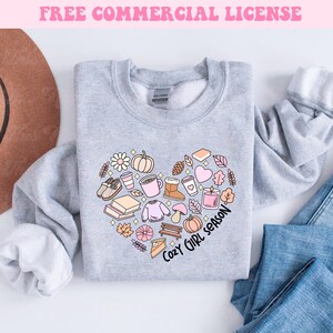 Op de afbeelding: Een grijs sweatshirt met een hartvormig ontwerp met verschillende herfstthema-illustraties, waaronder een pompoen, een mok, een boek en een trui. De tekst "Cozy Girl Season" staat onder het hart.