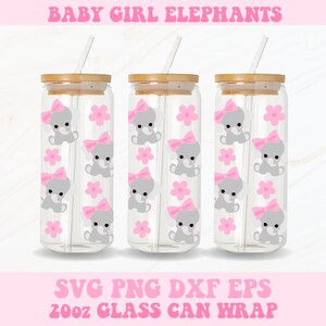 Puede incluir: Tres vasos de vidrio transparentes con tapas de bambú y pajitas. Cada vaso presenta un patrón repetido de flores rosas y elefantes bebés grises con lazos rosas. Los vasos están etiquetados con "20oz Glass Can Wrap".
