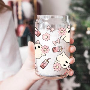 May include: A clear glass can-shaped cup with a white interior, decorated with pink and cream-colored ghost and flower graphics. The cup also features pink bows and drink illustrations. The cup is held by a person.