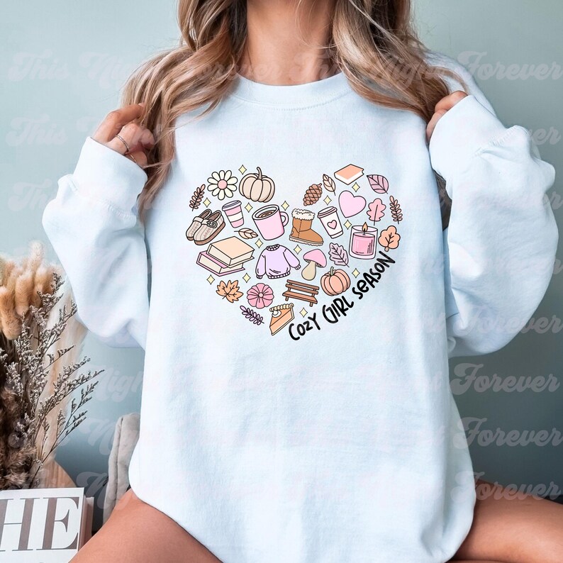 Op de afbeelding: Een lichtblauw sweatshirt met een hartvormig ontwerp met verschillende herfstthema-illustraties, waaronder pompoenen, bladeren, koffiebekers en een boek. De tekst "Cozy Girl Season" staat in het hart.