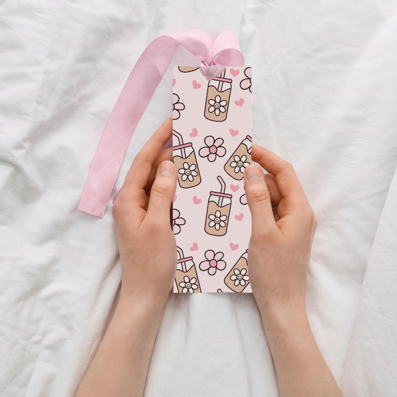 May include: A pink bookmark with a repeating pattern of iced coffee drinks with flower designs and pink hearts. The bookmark is held by hands and has a pink ribbon attached at the top.