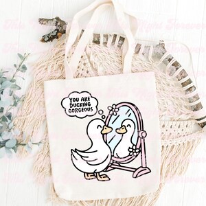 K&ouml;nnte beinhalten: Eine wei&szlig;e Canvas-Tragetasche mit einer rosa-wei&szlig;en Illustration eines Entchens, das sich in einem Spiegel betrachtet. Der Text "You are ducking gorgeous" befindet sich in einer Gedankenblase &uuml;ber dem Entchen.