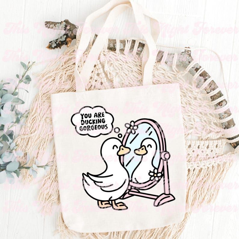 K&ouml;nnte beinhalten: Eine wei&szlig;e Canvas-Tragetasche mit einer rosa-wei&szlig;en Illustration eines Entchens, das sich in einem Spiegel betrachtet. Der Text "You are ducking gorgeous" befindet sich in einer Gedankenblase &uuml;ber dem Entchen.