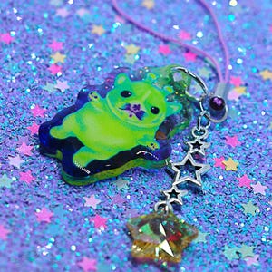 May include: A green cartoon dog charm with a star-shaped charm and a string. The charm has a purple and blue border. The background is a glittery purple surface with star-shaped confetti. The text "HamHamClubhouse" is at the bottom.