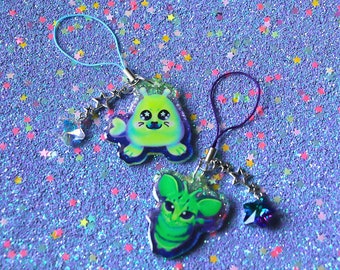 May include: Two colorful phone charms featuring cartoon alien characters. The charms are made of clear resin with glitter embedded inside. One charm is green with a blue and white alien with large eyes. The other charm is green and purple with a green alien with large eyes. Both charms have a silver chain and a star charm.