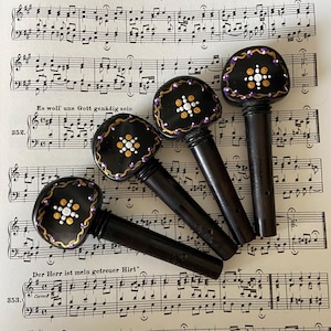 Può includere: Quattro piroli per violino neri con disegni dipinti in viola e oro. Ogni pirolo ha un design diverso con punti bianchi e un contorno viola.