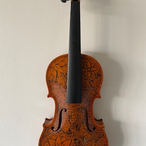 Puede incluir: Un violín marrón con un cuello negro y diseños intrincados de garabatos negros y blancos en el cuerpo. El diseño incluye flores, espirales y patrones geométricos.
