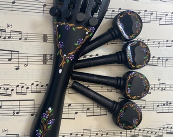 May include: Five black violin tuning pegs with floral designs. The pegs have a black finish and are decorated with gold accents and purple flowers. The pegs are arranged on a sheet of music.