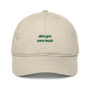 Może przedstawiać: Beżowa czapka z daszkiem z zielonym haftem z napisem "aloe you vera much".