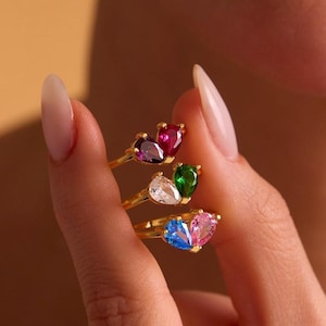 May include: A hand displays four gold rings, each with a teardrop-shaped gemstone in various colours: red, purple, green, white, blue, and pink. The rings have a delicate, minimalist design.