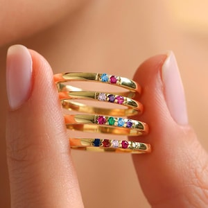 May include: A set of five gold rings with colorful gemstones. Each ring has a single gemstone in a different color, including red, blue, green, pink, and white.