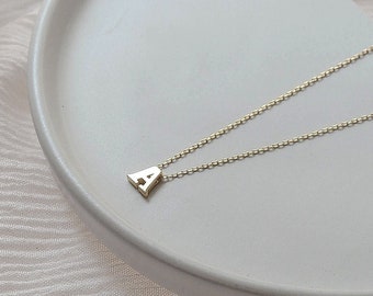 May include: A gold necklace with a small letter 'A' pendant on a white ceramic plate. The plate is on a white fabric background.