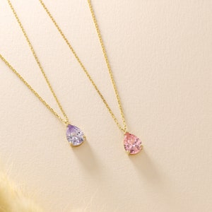 May include: Two gold necklaces, each featuring a teardrop-shaped gemstone pendant. One pendant is lavender, the other is pink. The pendants are set in gold and suspended from fine gold chains. The background is a soft, neutral colour.