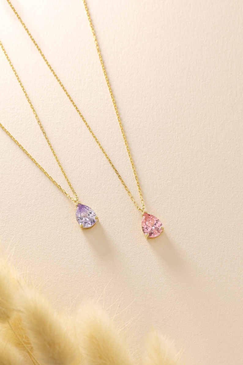 May include: Two gold necklaces, each featuring a teardrop-shaped gemstone pendant. One pendant is lavender, the other is pink. The pendants are set in gold and suspended from fine gold chains. The background is a soft, neutral colour.