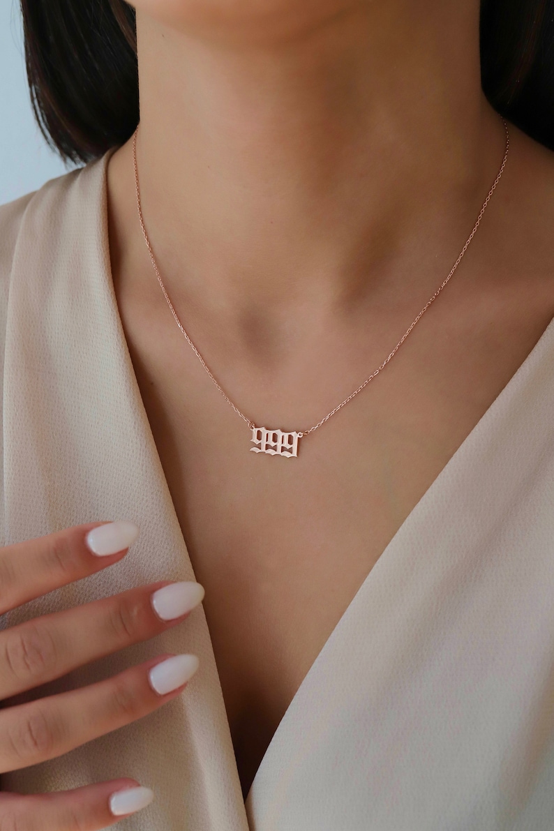 May include: A rose gold necklace with a delicate chain and a pendant featuring the number 1999 in a gothic font.