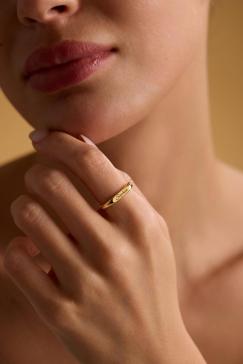 May include: A gold ring engraved with the name "Sophia". The ring has a simple band design, and the text is in a delicate, cursive font. The ring is worn on a finger.