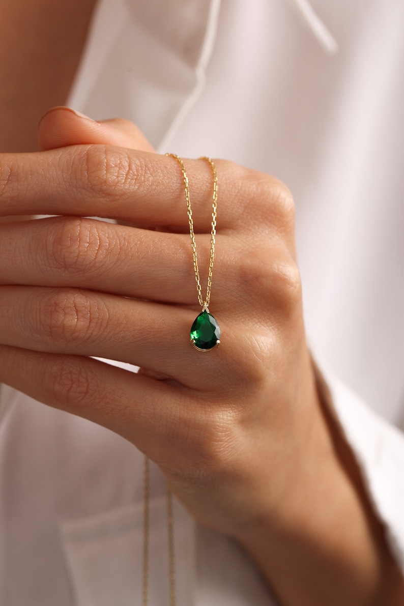May include: A gold necklace featuring a pear-shaped, emerald-green gemstone pendant. The pendant hangs from a fine gold chain. The necklace is shown against a white backdrop.