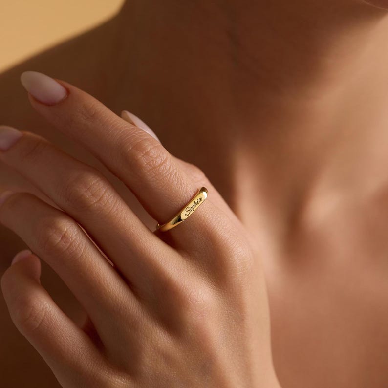 May include: A gold ring engraved with the name "Sophia". The ring is worn on a finger, highlighting its simple, elegant design. The ring appears to be made of polished metal, reflecting light. The ring's design is minimalist.