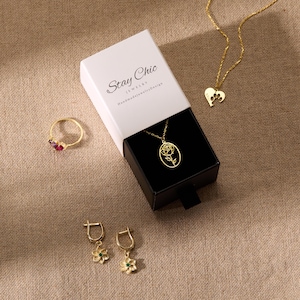 Puede incluir: Una variedad de joyas doradas, que incluyen un anillo con piedras rojas, un collar con un colgante de rosa y un par de pendientes de flores con detalles verdes. Tambi&eacute;n es visible un collar con un colgante de coraz&oacute;n. Las joyas se exhiben con una caja de regalo.