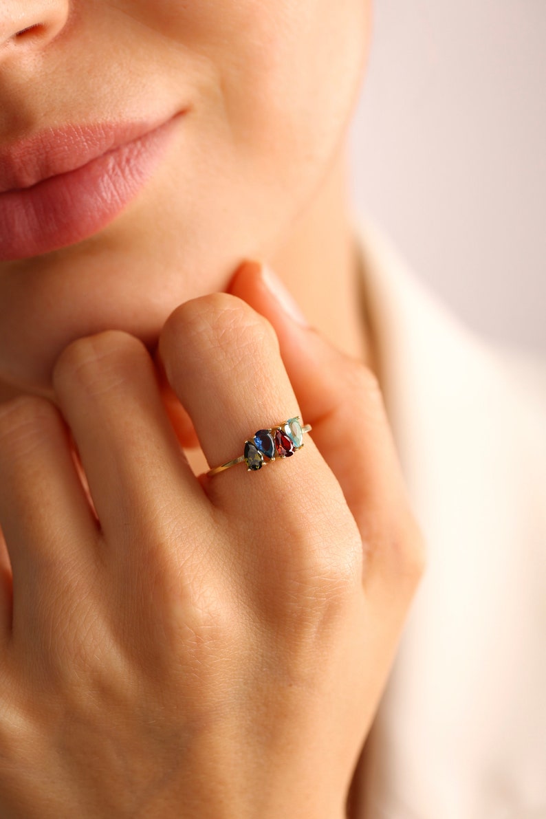 May include: A gold ring with four colorful gemstones. The gemstones are blue, green, red, and blue. The ring is on a person's finger.