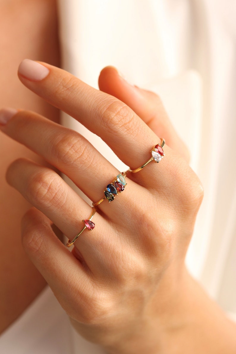 May include: A hand wearing four gold rings with colorful gemstones. The rings feature pear-shaped stones in shades of red, blue, green, and white.