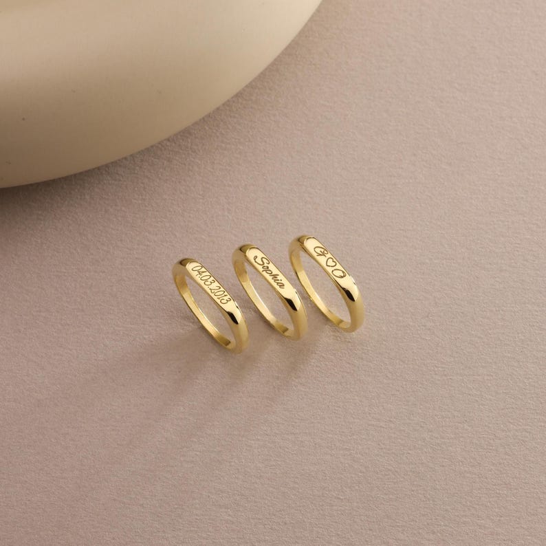 May include: Three gold rings with engraved inscriptions. One ring displays "04.03.2018", another reads "Sophia", and the third features a heart symbol and the letters "GOO". The rings are presented on a neutral-toned surface.