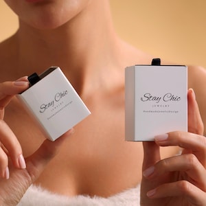 May include: Two white jewellery boxes with black ribbon closures are held up. The boxes have the words "Stay Chic" and "Jewelry HandmadeJewelryDesign" printed on them in black. The background is a solid tan colour.