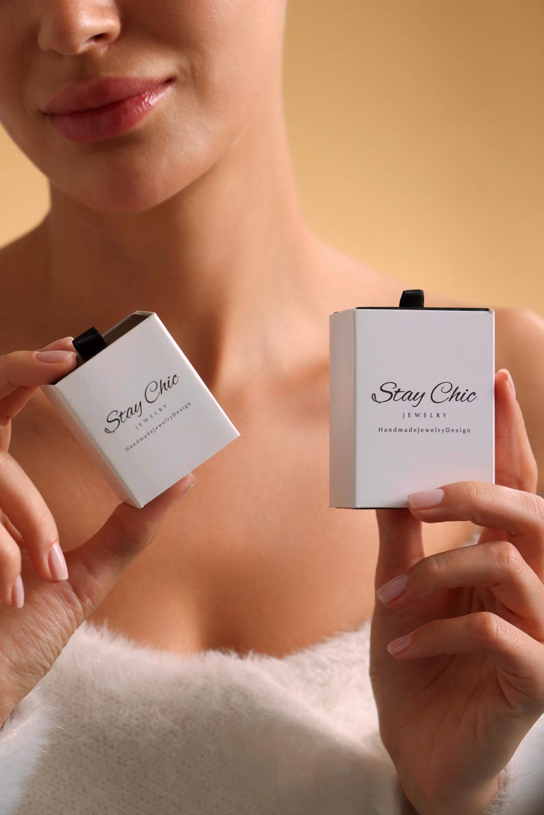 May include: Two white jewellery boxes with black ribbon closures are held up. The boxes have the words "Stay Chic" and "Jewelry HandmadeJewelryDesign" printed on them in black. The background is a solid tan colour.