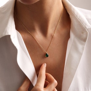 May include: A gold necklace featuring a teardrop-shaped, emerald-green gemstone pendant. The necklace is worn against a white collared shirt. The pendant is approximately 1.3 cm in length.