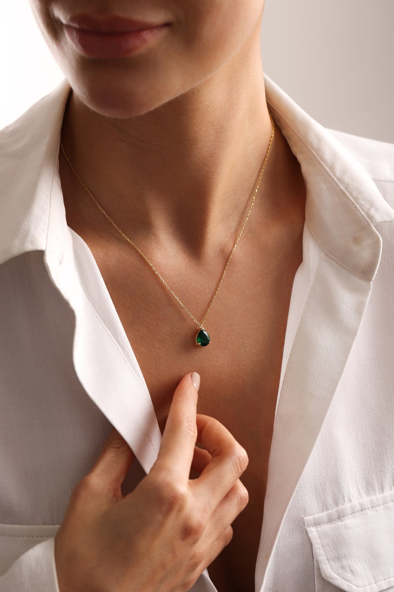 May include: A gold necklace featuring a teardrop-shaped, emerald-green gemstone pendant. The necklace is worn against a white collared shirt. The pendant is approximately 1.3 cm in length.