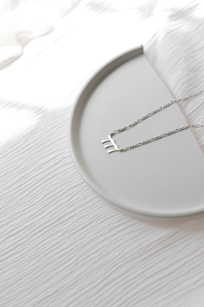 May include: A silver necklace with a simple, abstract pendant on a white background.