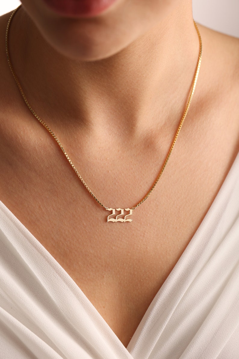 May include: A gold chain necklace with a pendant featuring the number 222 in a gothic font.