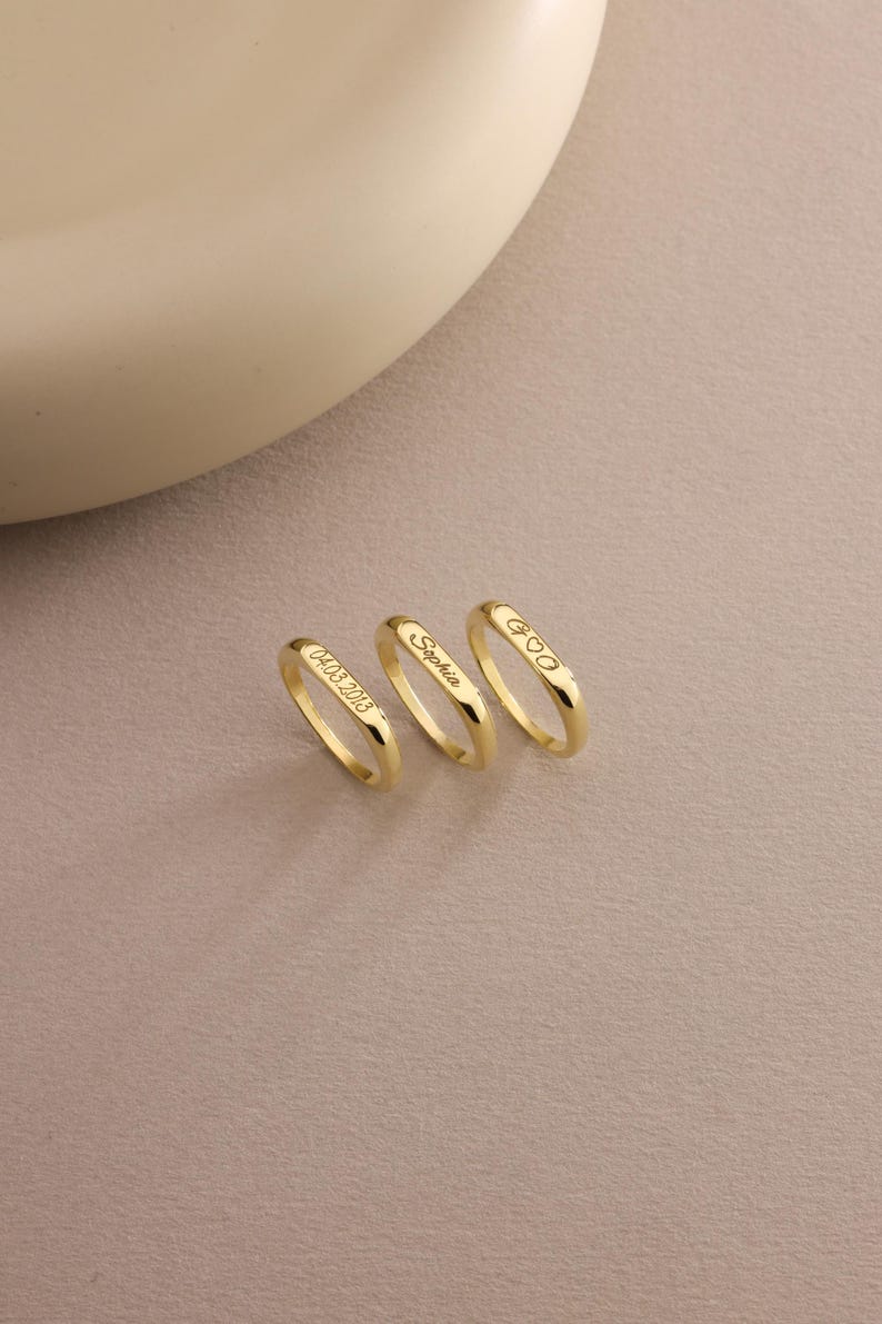 May include: Three gold rings with engraved inscriptions. One ring displays the date "04.03.2018", another the name "Sophia", and the third the word "Goo". The rings are presented on a neutral-toned surface.