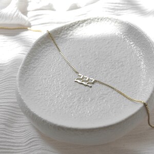 May include: A gold chain necklace with a delicate, wave-shaped pendant. The necklace is displayed on a white ceramic dish.