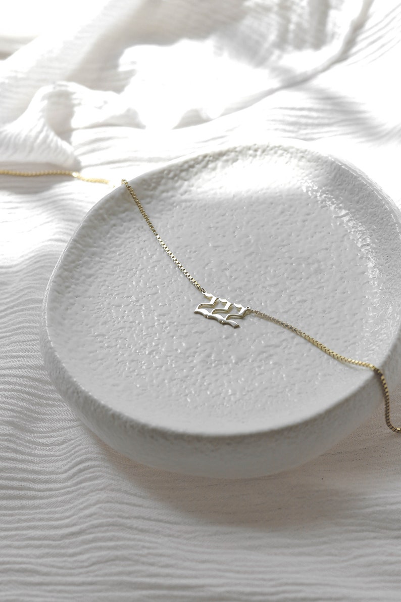 May include: A gold chain necklace with a delicate, wave-shaped pendant. The necklace is displayed on a white ceramic dish.