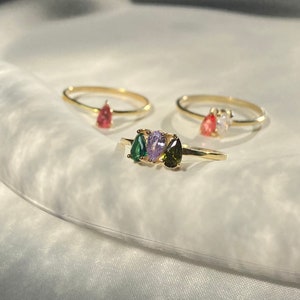 May include: Three gold rings with different colored gemstones. The rings feature a pear-shaped green stone, a pear-shaped purple stone, and a pear-shaped red stone.