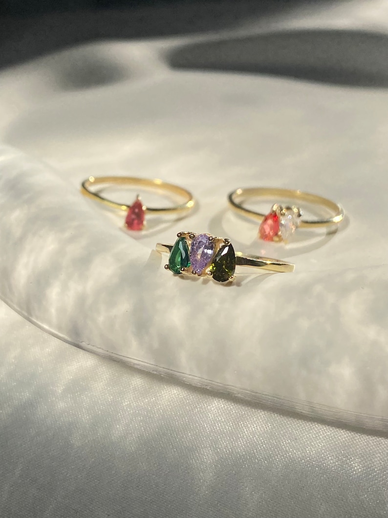 May include: Three gold rings with different colored gemstones. The rings feature a pear-shaped green stone, a pear-shaped purple stone, and a pear-shaped red stone.