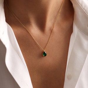 May include: A gold necklace featuring a dark green, teardrop-shaped gemstone pendant. The necklace is displayed against a white collared shirt.