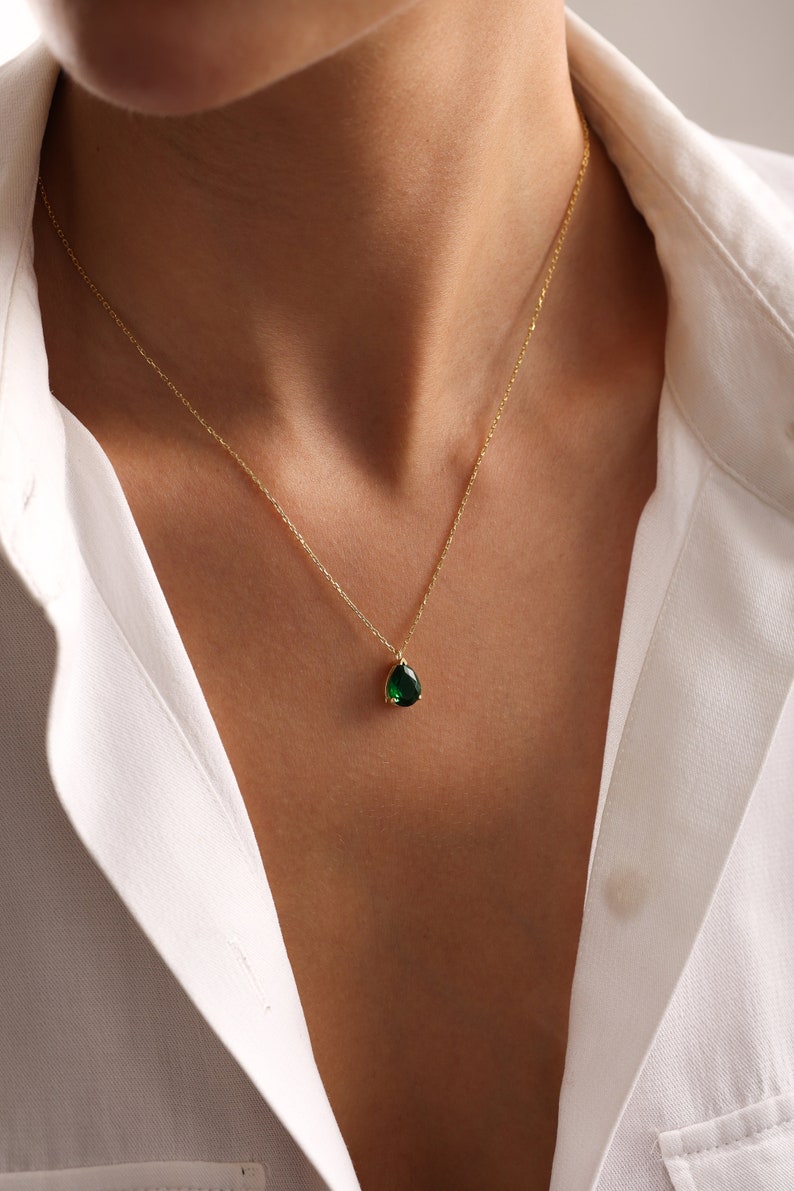 May include: A gold necklace featuring a dark green, teardrop-shaped gemstone pendant. The necklace is displayed against a white collared shirt.