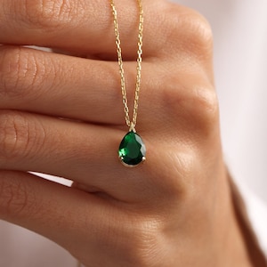 May include: A gold necklace with a pear-shaped, emerald-green gemstone pendant. The pendant hangs from a delicate gold chain. The necklace is shown against a plain background, emphasising the vibrant green of the gemstone.