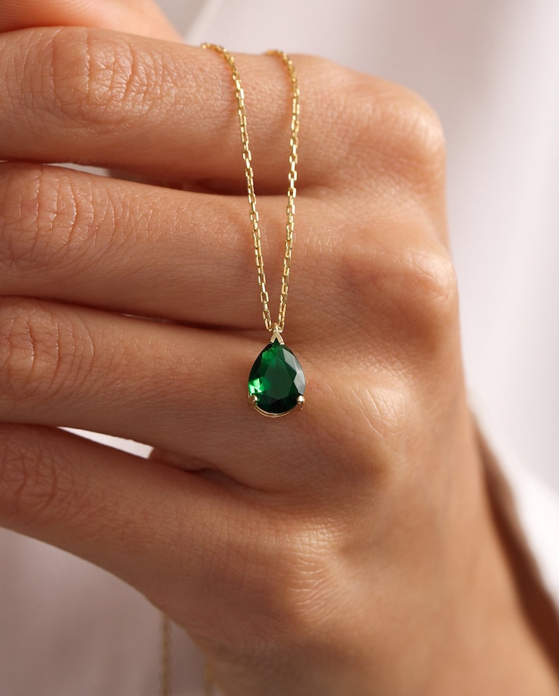 May include: A gold necklace with a pear-shaped, emerald-green gemstone pendant. The pendant hangs from a delicate gold chain. The necklace is shown against a plain background, emphasising the vibrant green of the gemstone.