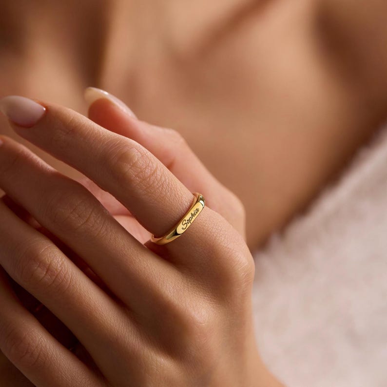 May include: A gold ring with the name "Sophia" inscribed on the band. The ring is displayed on a finger, emphasizing its minimalist design. The polished surface of the ring reflects light, accentuating the engraved name.