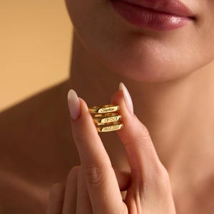 May include: Four gold rings, each engraved with text: "Sophia", "GOO", and "04.03.2013". The rings are held between fingers. The background is a soft, neutral tone.