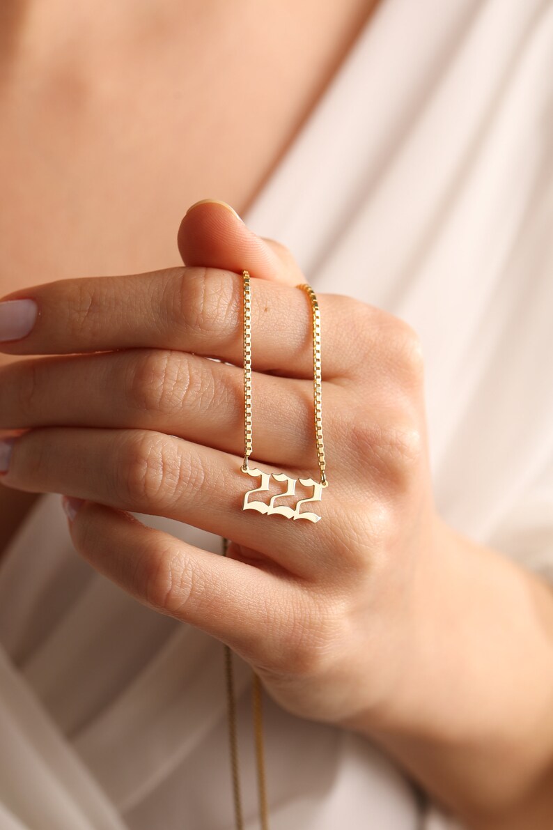 May include: A gold chain necklace with a pendant featuring the number 222 in a gothic font.