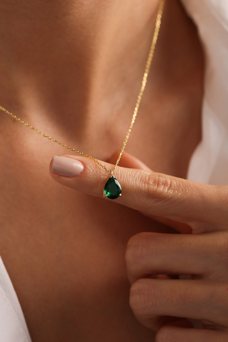 May include: A gold necklace with a teardrop-shaped, emerald-green gemstone pendant. The pendant hangs from a fine gold chain. The necklace is held up by a person's fingers. The necklace is a piece of jewellery.