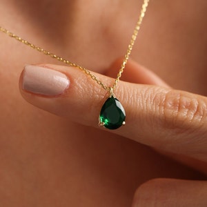 May include: A gold necklace with a pear-shaped, emerald-green gemstone pendant. The pendant hangs from a fine gold chain. The necklace is shown against a blurred background.