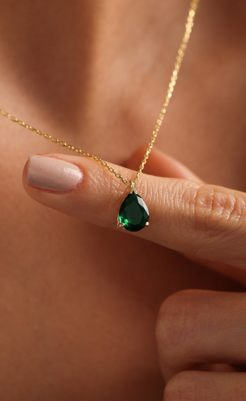 May include: A gold necklace with a pear-shaped, emerald-green gemstone pendant. The pendant hangs from a fine gold chain. The necklace is shown against a blurred background.