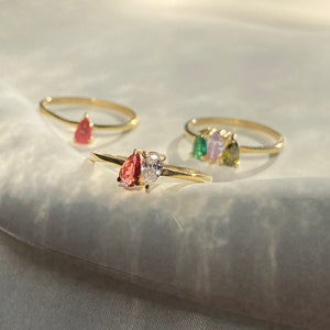 May include: Three gold rings with pear-shaped gemstones. The first ring has a red gemstone, the second ring has a clear gemstone, and the third ring has three gemstones: green, purple, and green.